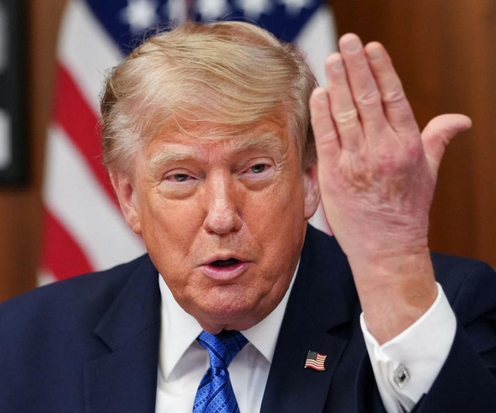 Donald Trump z uniesioną dłonią na tle flagi USA. O napięciu na linii Waszyngton-Teheran przeczytasz w Eska.
