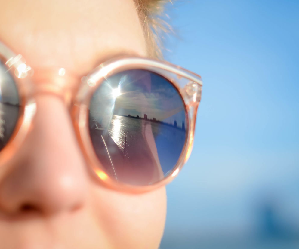 Zbliżenie na twarz osoby w beżowych okularach przeciwsłonecznych, w których odbija się błękitne niebo z promieniami słońca i odległa panorama miasta nad wodą. Okulary bez odpowiedniego filtra UV mogą szkodzić oczom bardziej niż ich brak, o czym przeczytasz na Poradnik Zdrowie.