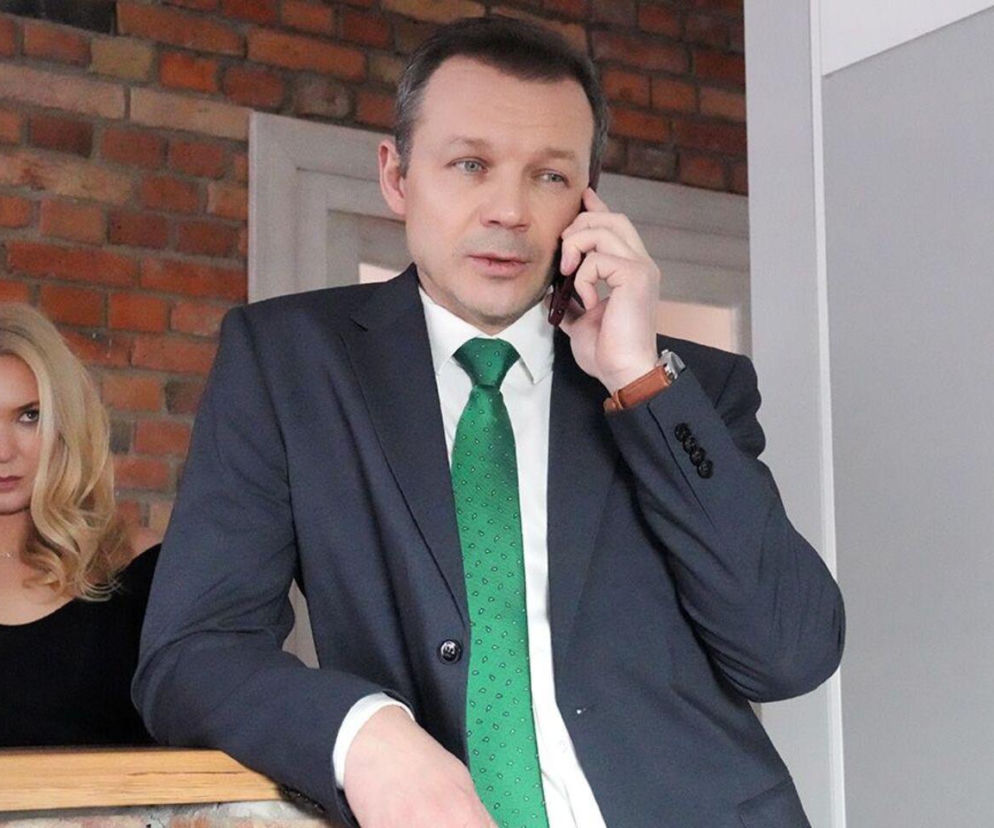 Lesław Żurek jako Bruno z serialu Barwy szczęścia rozmawia przez telefon, opierając się o drewnianą balustradę. W tle, z lewej strony, widoczna jest Marta Dąbrowska jako Karolina z niezadowoloną miną. Zdjęcie ilustruje intrygi postaci, o których można przeczytać na Super Seriale.