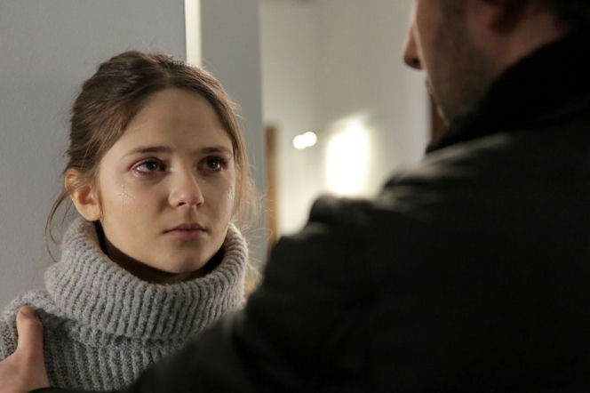 Monika Mielnicka jako Lilka z serialu M jak miłość ze łzami w oczach, w szarym golfie, trzymana za ramię przez mężczyznę w ciemnej kurtce. Emocjonalna scena z dramatycznego odcinka 1515, którą można zobaczyć na Super Seriale.