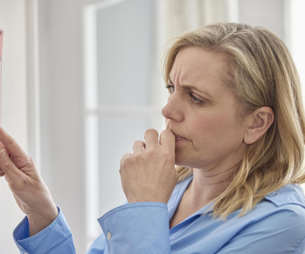 Zamyślona kobieta w jasnoniebieskiej koszuli z blond włosami, dotyka palcem kalendarza ściennego, z drugą dłonią przy ustach, co symbolizuje zapominanie. Obraz odzwierciedla problem cyfrowej amnezji i jest częścią artykułu na Poradnik Zdrowie o tym, jak smartfony wpływają na pamięć.