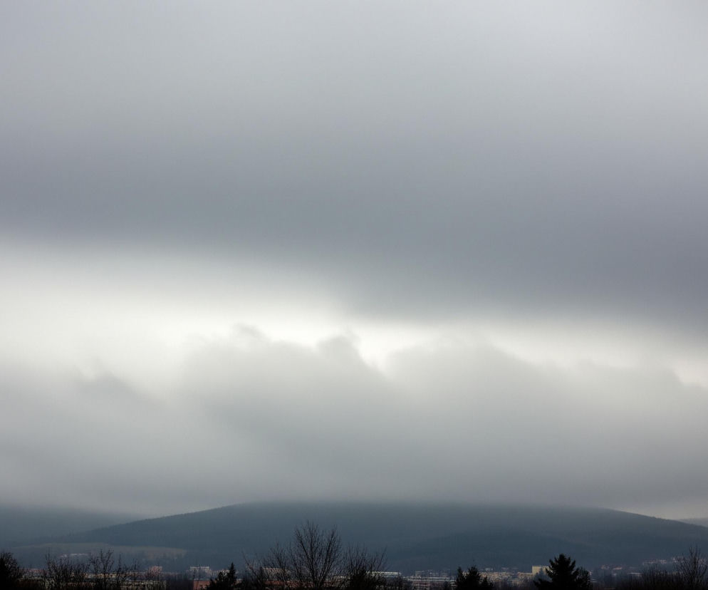 Widok z lotu ptaka przedstawia mglisty, pochmurny krajobraz z ciemnymi pagórkami lub górami w tle, częściowo zakrytymi niskimi chmurami. W dolnej części obrazu widoczne są fragmenty zabudowań miejskich oraz korony nagich drzew, a niebo zajmuje większą część kadru. Kolorystyka utrzymana jest w szarościach, od jasnych, niemal białych partii chmur, po ciemniejsze, niemal granatowe odcienie pasm górskich.
