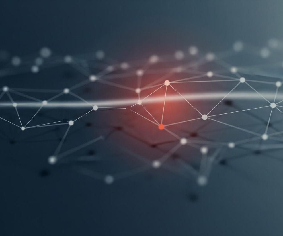 Apstraktne trójwymiarowe odwzorowanie sieci połączeń z licznymi węzłami i liniami wypełnia centralną część obrazu. Białe kropki, symbolizujące węzły, są połączone cienkimi, białymi liniami, tworząc wielokątne struktury. Kilka węzłów w centralnym obszarze jest podświetlonych na czerwono, co sugeruje punkt skupienia, otoczony rozmyciem i czerwonym blaskiem. Całość kompozycji ma ciemnoszare tło, które przechodzi w jaśniejsze odcienie szarości w górnej części, natomiast biała, pozioma smuga światła przecina obraz w okolicy środka.