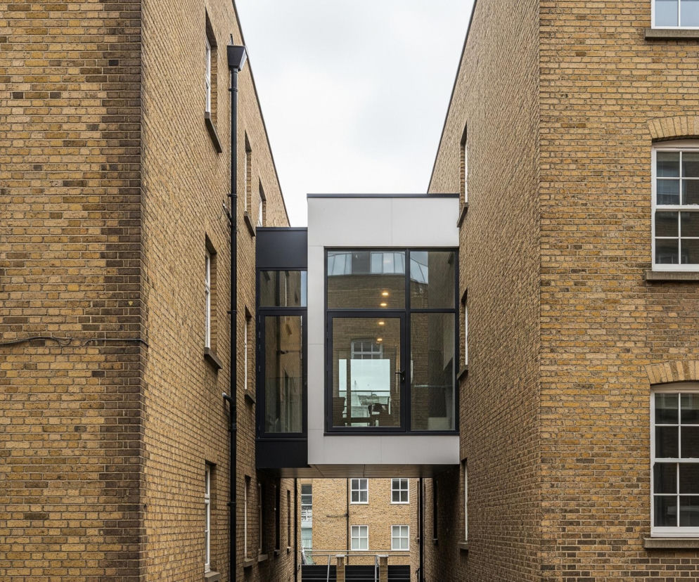 Dwa ceglane budynki w odcieniach piaskowego brązu i beżu tworzą wąską szczelinę, na której wysokości środkowej części znajduje się współczesne, przeszklone przejście. Przejście to ma białe i czarne akcenty, a w jego wnętrzu widać odbijające się elementy, w tym okna przeciwległych budynków. Na ceglanych fasadach rozmieszczone są prostokątne, białe okna z podziałami, a na prawej ścianie widoczna jest czarna rura spustowa biegnąca wzdłuż budynku. Pomiędzy budynkami, w tle, widoczne są fragmenty innych ceglanych konstrukcji z oknami, a w górnej części dominuje jasne, zachmurzone niebo.