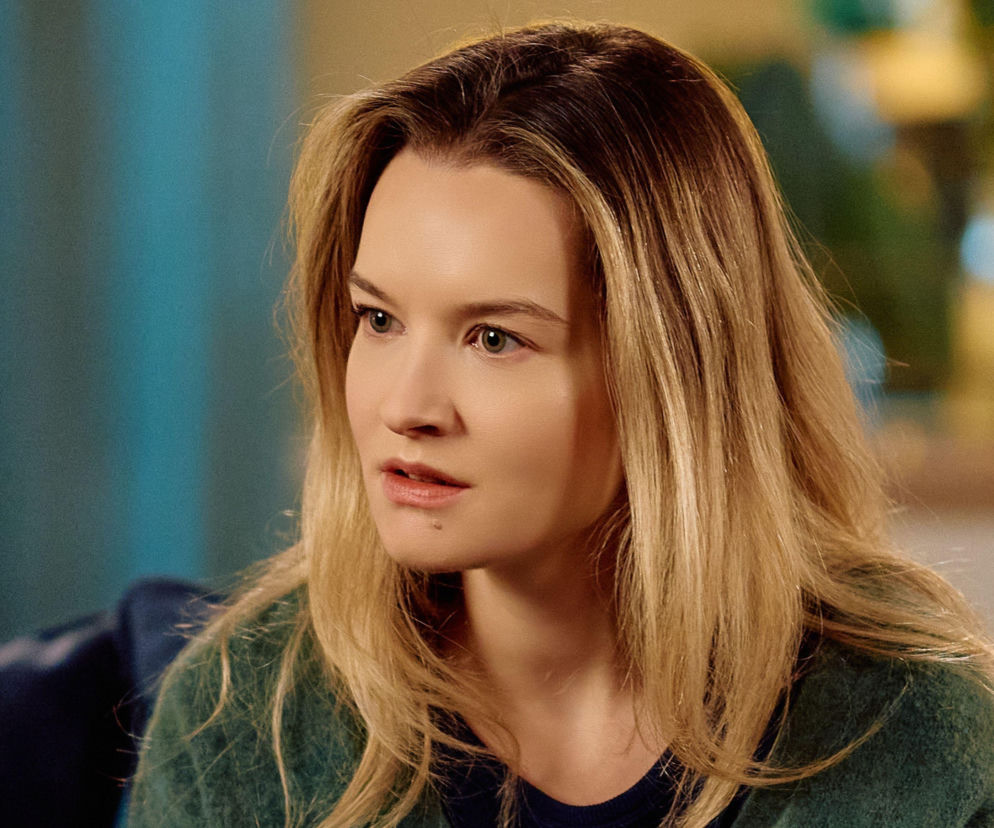 Katarzyna Gałązka jako Kalina z serialu Na Wspólnej zaniepokojona odkryciem Darka, w zbliżeniu, z jasnymi włosami i w ciemnozielonym swetrze, z widocznym błękitnym tłem. O kulisach serialu i innych Super Serialach przeczytasz na naszym portalu.