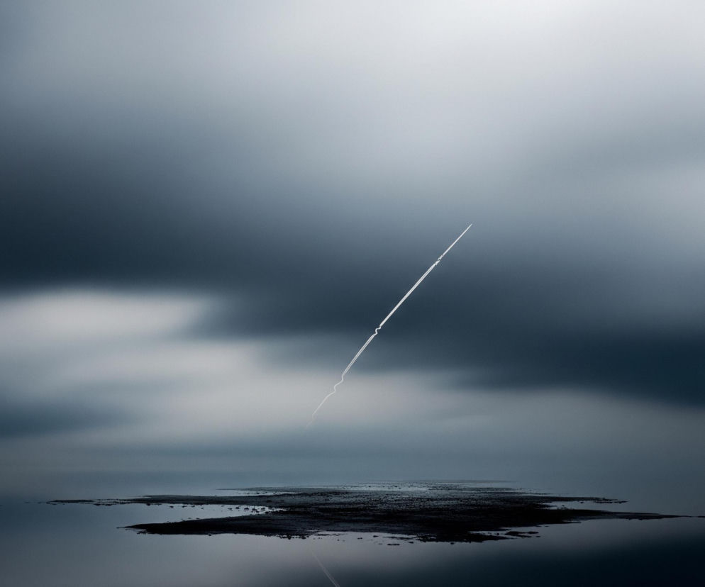 Na obrazie dominuje mroczne, zamglone niebo w odcieniach szarości i granatu, z jaśniejszym obszarem w górnym prawym rogu. Przez środek kadru, pod kątem, przecina smuga kondensacyjna samolotu, tworząc białą linię, która lekko zanika w dolnej części. Poniżej rozciąga się ciemny horyzont z plamą nierównego, ciemnego lądu lub płycizny, odzwierciedlającego częściowo smugę na powierzchni wody lub wilgotnej ziemi.