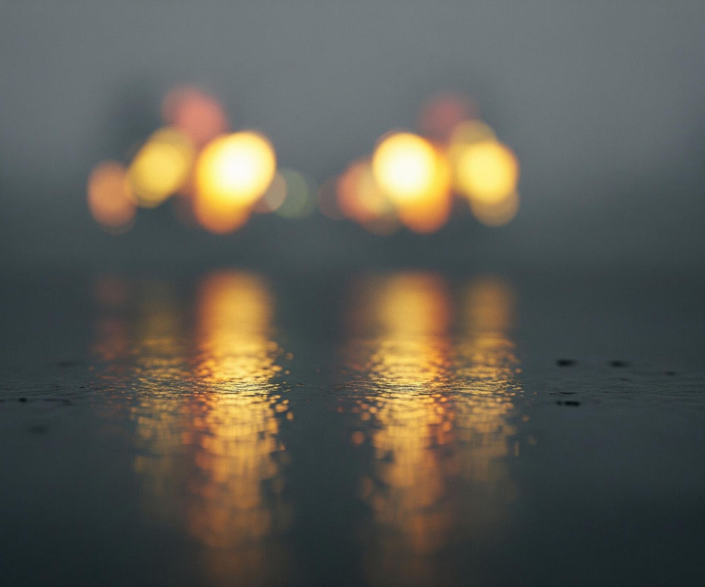 Centralna część obrazu przedstawia rozmyte, intensywnie żółte i pomarańczowe światła odbijające się w mokrej, ciemnej powierzchni. Na horyzoncie widoczne są dwie główne, owalne plamy jasnego światła, z pomniejszymi, mniej wyraźnymi plamami po bokach, wszystkie rozmyte w efekcie bokeh. Poniżej, na pierwszym planie, te same światła odbijają się w falującej powierzchni, tworząc jaśniejsze, bardziej rozciągnięte smugi koloru złotego i pomarańczowego. Całość tonie w mrocznym, szarawym tle, sugerującym mgłę lub zmierzch.