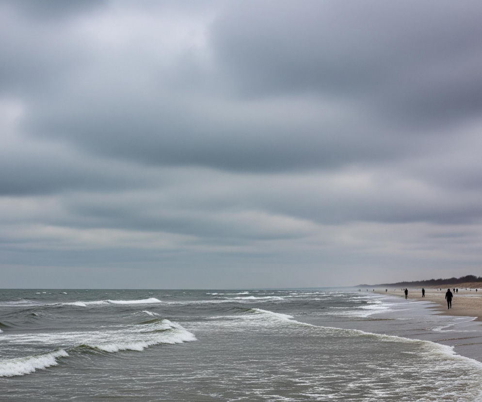 Szary ocean z licznymi falami wznosi się i opada, tworząc białe grzywy z piany, które rozbijają się o piaszczysty brzeg, częściowo mokry od wody. Na plaży, w oddali, idzie kilka osób ubranych w ciemne kurtki, a jedna z nich nosi białą czapkę. Nad całą sceną rozciąga się zachmurzone, szare niebo, dominujące nad horyzontem.