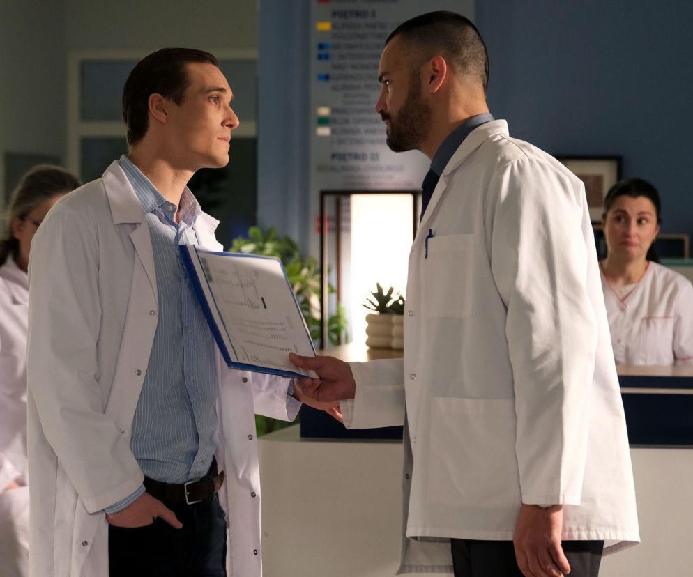 Maurycy Popiel jako Olek i Mikołaj Krawczyk jako Sowiński w białych fartuchach lekarzy, rozmawiający ze sobą na korytarzu szpitala, gdzie Sowiński wręcza Olkowi teczkę. Scena z serialu M jak miłość. Więcej o tym wątku na Super Seriale.