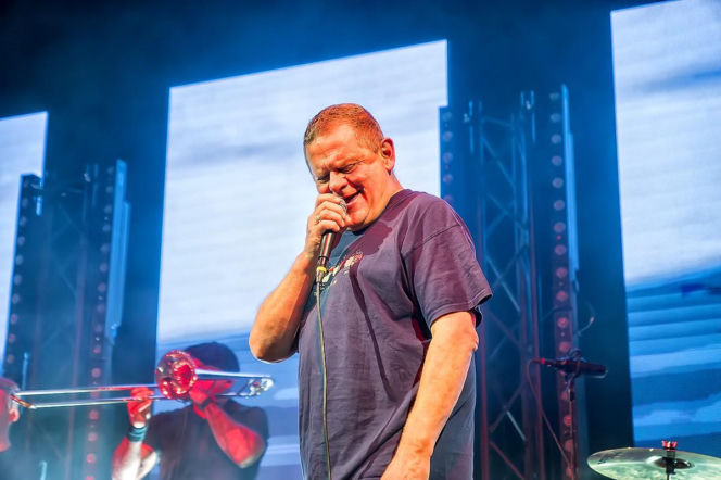 Kazik Staszewski, lider zespołu Kult, śpiewający na scenie z mikrofonem w dłoni, z widoczną ekspresją na twarzy, podczas jednego z koncertów. W tle zarysy muzyków i sprzętu scenicznego. Więcej o stanie zdrowia artysty przeczytasz na portalu Poradnik Zdrowie.