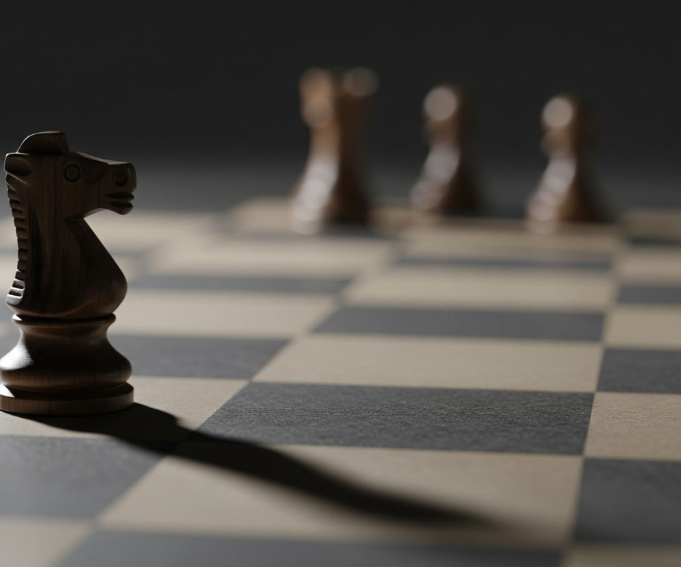 Ciemnobrązowy skoczek szachowy stoi na jasnym polu szachownicy w lewej części kadru. Od figury na ukos w prawo rozciąga się długi, ostry cień. Tło przedstawia rozmazane figury szachowe w kolorze jasnobrązowym, ustawione w rzędzie. Całość jest słabo oświetlona, z punktowym światłem padającym na skoczka, podkreślając jego krawędzie i rzucając kontrastujący cień na szachownicę.