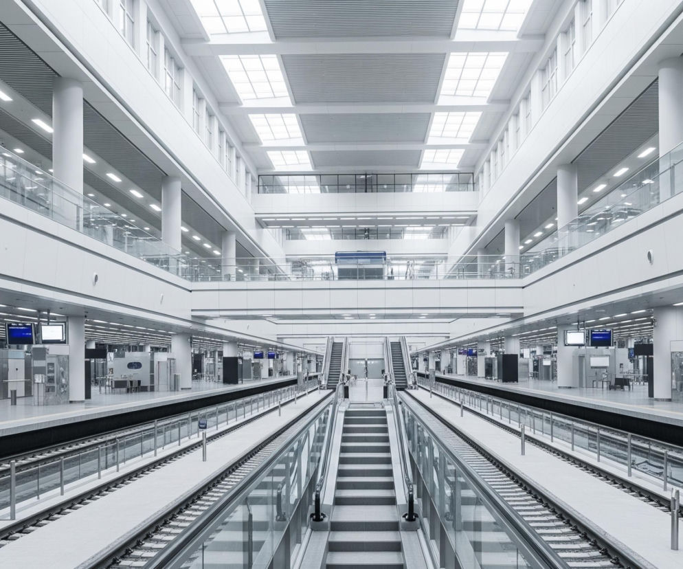 Nowoczesna hala dworca kolejowego lub metra, utrzymana w jasnej, monochromatycznej kolorystyce, dominuje w centrum kadru. Na pierwszym planie widoczne są dwa tory kolejowe oddzielone wąskim, jasnym pasem. Po obu stronach torów rozciągają się szerokie perony z licznymi filarami, ekranami informacyjnymi i punktami usługowymi, na których nie widać żadnych ludzi. W tle, powyżej peronów, znajdują się kolejne poziomy budynku z przeszklonymi balustradami, a sufit jest częściowo przeszklony, wpuszczając naturalne światło i uzupełniony o rzędy jasnego, sztucznego oświetlenia.