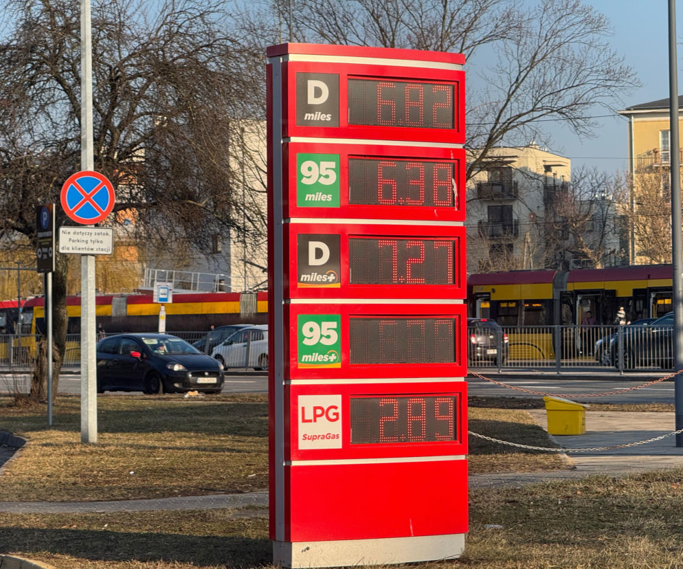 Czerwony pylon z aktualnymi cenami paliw na stacji benzynowej: Diesel (D) za 6,82 zł/l, Benzyna 95 za 6,38 zł/l i LPG za 2,89 zł/l. W tle ruch uliczny z tramwajem i samochodami, odzwierciedlający rosnące koszty dla kierowców, o czym możesz przeczytać w Super Biznes.