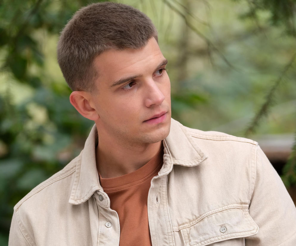 Młody mężczyzna, Mateusz (Rafał Kowalski), z krótkimi włosami i w beżowej koszuli, spogląda w prawą stronę, z zamyśloną miną na tle rozmazanej zieleni. Informacje o wzruszających zaręczynach Mateusza w M jak miłość znajdziesz na Super Seriale.