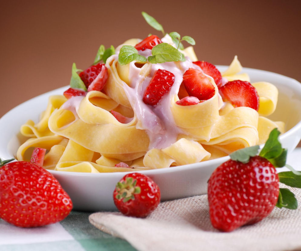 Porcja makaronu tagliatelle z truskawkami, jogurtem i listkami mięty w białej misce na tle obrusu w zieloną kratkę, z truskawkami leżącymi obok. Sprawdź na Poradnik Zdrowie, czy cukrzyk może jeść makaron i jaki wybrać.