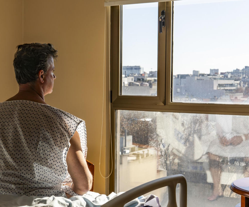 Pacjent w szpitalnej koszuli siedzący na łóżku i patrzący przez okno, zamyślony o przyszłości i czekający na diagnozę nowotworu. Artykuł na Poradnik Zdrowie podkreśla dramatyczne opóźnienia w polskim systemie opieki zdrowotnej.