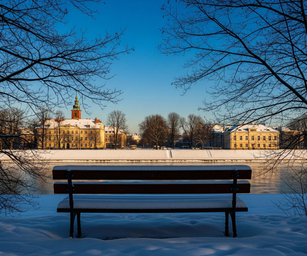 Centralnie na pierwszym planie znajduje się drewniana ławka z metalowymi nogami, cała pokryta warstwą białego śniegu. W tle rozciąga się rzeka, której powierzchnia odbija błękitne niebo i złociste światło zachodzącego słońca, tworząc jasną, poziomą linię. Po obu stronach rzeki, na horyzoncie, widać szereg budynków w ciepłych, żółtych i pomarańczowych odcieniach, z zielonym dachem wieży kościelnej po lewej stronie. Cała scena jest otoczona drzewami bez liści, ich ciemne gałęzie tworzą naturalną ramę obrazu na tle jasnoniebieskiego nieba.