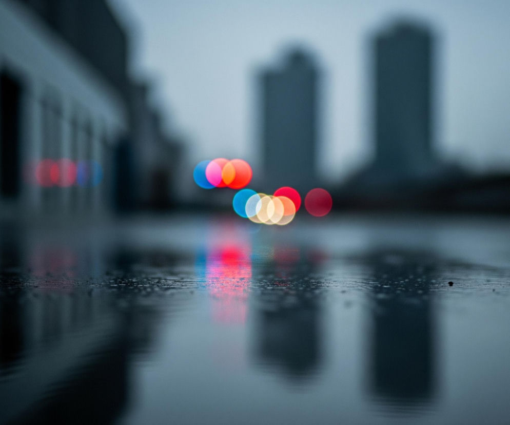 Mokra, odblaskowa nawierzchnia zajmuje dolną połowę obrazu, z wyraźnie widocznymi kroplami wody. Powyżej linii horyzontu, na środku kadru, rozmywają się jaskrawe, okrągłe światła bokeh w kolorach czerwonym, niebieskim, różowym, pomarańczowym i żółtym. Po lewej stronie w tle widoczne są niewyraźne, pionowe kształty budynków w odcieniach szarości, a po prawej, na tle zamglonego, jasnoszarego nieba, rysują się dwie smukłe, ciemniejsze bryły wieżowców. Kolorowe światła odbijają się w wilgotnej nawierzchni, tworząc podłużne, rozmazane refleksy.
