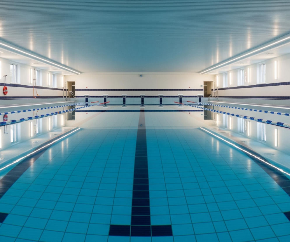 Basen pływacki o krystalicznie czystej, błękitnej wodzie zajmuje centralną część kadru, z dwiema linami torowymi w czerwono-białych i niebiesko-białych barwach rozciągającymi się wzdłuż krawędzi, dzielącymi go na tory. Na ścianach wzdłuż basenu widoczne są białe płytki z ciemnoniebieskimi pasami dekoracyjnymi oraz prostokątne okna w regularnych odstępach, a także pionowe świetlówki, których odbicia w tafli wody tworzą świetliste, falujące smugi. U szczytu basenu znajdują się szare bloki startowe oraz drabinki wejściowe, a na lewej ścianie wisi pomarańczowe koło ratunkowe. Sufit jest pokryty poziomymi, jasnoniebieskimi panelami, z długimi, liniowymi lampami LED biegnącymi wzdłuż niego.