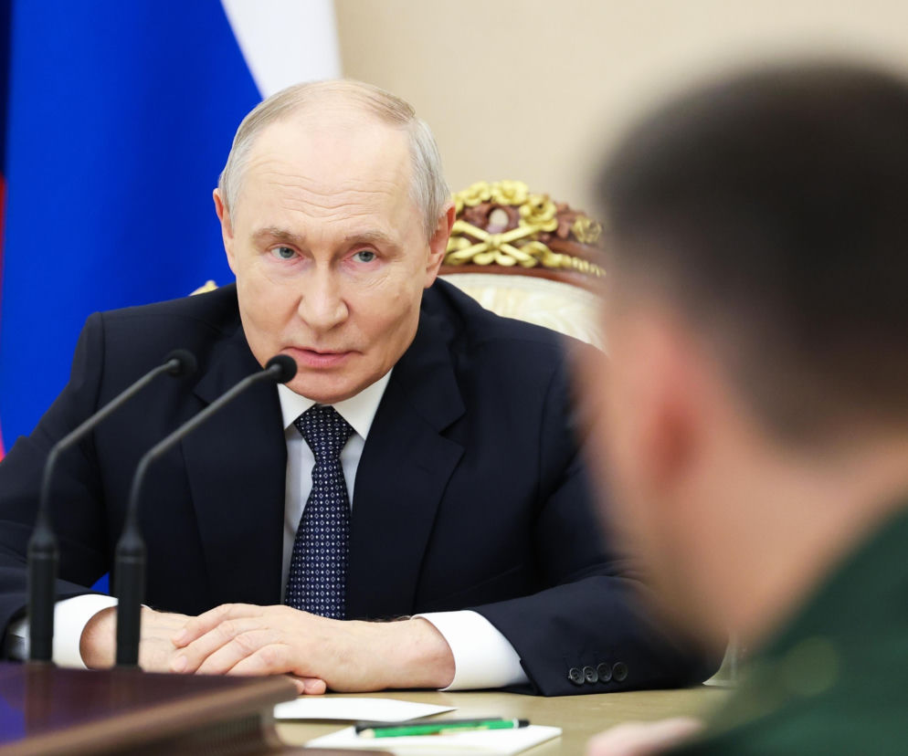 Władimir Putin siedzi przy stole z mikrofonami na tle rosyjskiej flagi. O groźbach Rosji wobec Londynu przeczytasz w Eska.