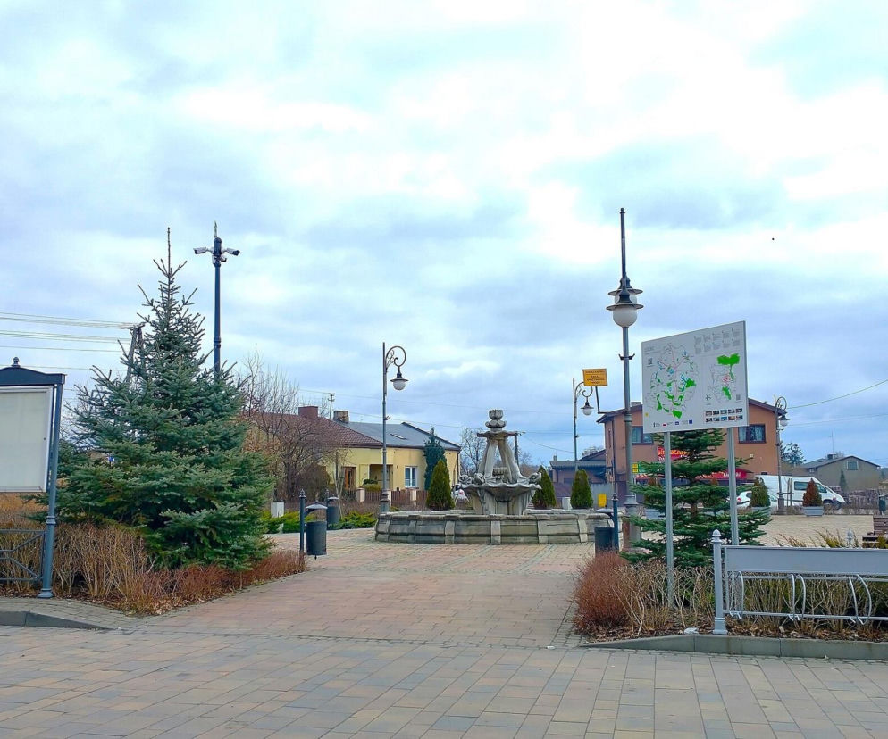 Plac w centrum Ujazdu z fontanną, latarniami i tablicą informacyjną. O historii miasta przeczytasz w Eska Łódź.
