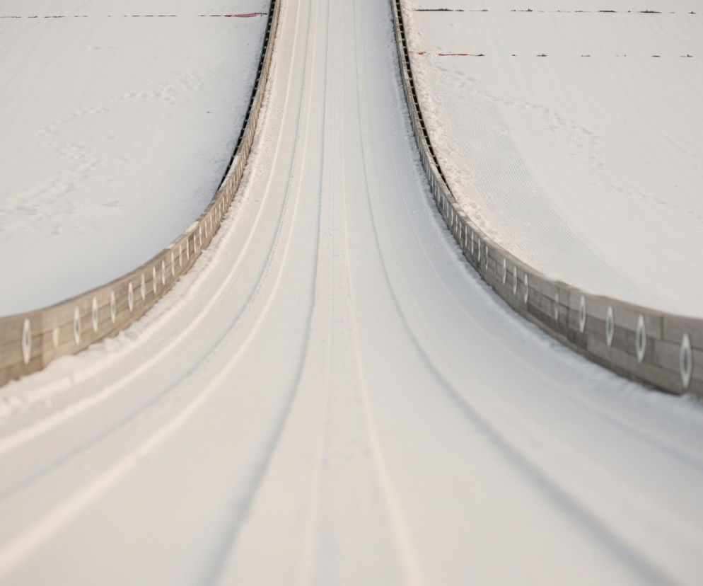 Głównym elementem obrazu jest pokryty śniegiem tor narciarski do skoków, biegnący centralnie w kierunku oddalenia. Tor ten składa się z dwóch równoległych, lekko wklęsłych rynien, oddzielonych wąskim, płaskim pasem, wszystko pokryte jest świeżym, białym śniegiem, na którym widoczne są drobne nierówności i ślady. Po obu stronach toru znajdują się drewniane barierki w kolorze brązowoszarym, z regularnie rozmieszczonymi jasnymi, owalnymi elementami. Za barierkami rozciągają się szerokie, ośnieżone stoki, na których gdzieniegdzie widać ciemne, drobne punkty, być może ślady roślinności lub niedoskonałości śniegu, oraz pojedyncze, delikatne ślady stóp. Kolorystyka obrazu to głównie biel śniegu z subtelnymi odcieniami szarości i beżu w drewnie, oświetlenie wydaje się być naturalne, tworząc delikatne cienie i podkreślając teksturę powierzchni.
