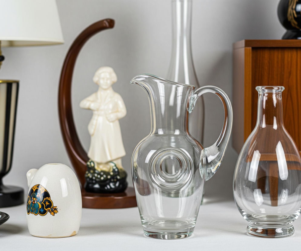 Przezroczysty szklany dzbanek z uchwytem i wyraźnym okrągłym zagłębieniem na środku stoi na białym blacie, dominując centralną część obrazu. Na prawo od dzbanka znajduje się mniejsza, również przezroczysta szklana butelka z szerokim dnem i zwężającą się szyjką, a za nią, po prawej stronie tła, widać drewnianą szafkę z widocznym usłojeniem. Po lewej stronie dzbanka stoi biała, jajowata figurka z barwnym, stylizowanym wzorem w odcieniach niebieskiego, żółtego i czarnego, natomiast za nią widoczna jest kremowa ceramiczna figurka człowieka umieszczona na ciemnobrązowej podstawie, która z kolei znajduje się na zaokrąglonym, drewnianym elemencie. Dalej w lewo, na blacie, leży mały, czarny talerzyk z niebiesko-białym wzorem kwiatowym, a za nim stoi lampa stołowa z biało-czarną, pionowo pasowaną podstawą i białym abażurem.