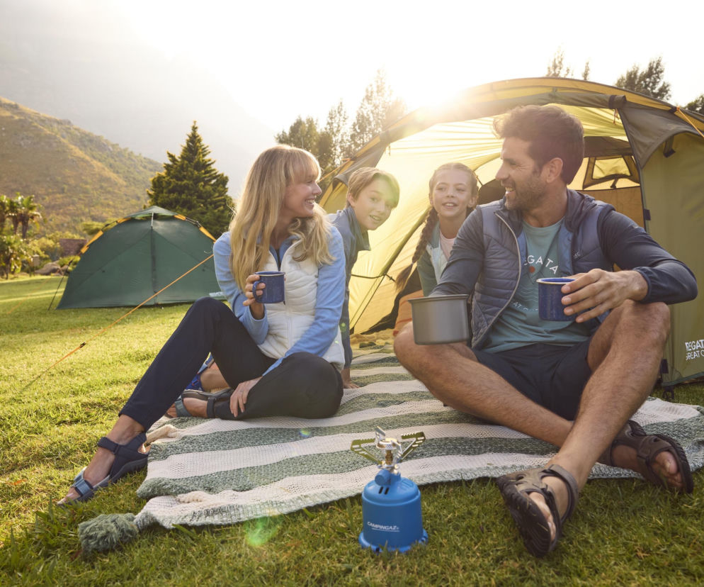 Uśmiechnięta rodzina siedząca na kocu przed namiotem marki Regatta Great Outdoors na tle górskiego krajobrazu, delektująca się poranną kawą na campingu. Obok nich turystyczna kuchenka gazowa. Na naszym portalu przeczytasz o produktach Regatta, idealnych na każdą wyprawę.