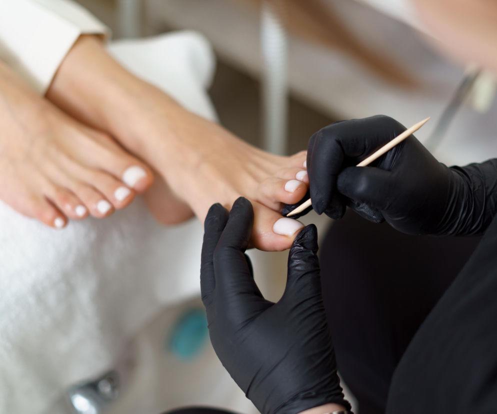Ręce kosmetyczki w czarnych rękawiczkach wykonującej pedicure. O kontrolach ZUS na L4 przeczytasz na SE Białystok.