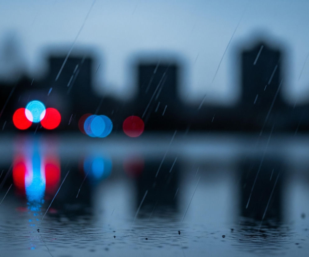 Przedni plan wypełnia powierzchnia wody z widocznymi, drobnymi kroplami deszczu rozbijającymi się o jej taflę, tworzącymi małe kręgi i fale. Nad wodą, w średnim planie, unosi się rozmyta panorama miasta z niewyraźnymi sylwetkami wysokich budynków na tle jasnoszarego, pochmurnego nieba. Wśród tych niewyraźnych kształtów, cztery wyraźnie świecące, rozmyte światła – dwa czerwone i dwa niebieskie – odbijają się w wodzie, tworząc smugi w tych samych barwach. Całość obrazu jest ciemna i zdominowana przez odcienie szarości i błękitu, z widocznymi, ukośnymi smugami symbolizującymi padający deszcz.