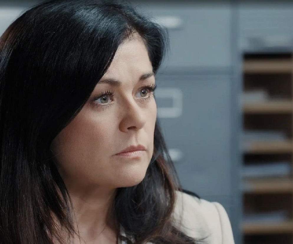 Katarzyna Cichopek jako Kinga Zduńska z serialu M jak miłość na przesłuchaniu, z poważnym wyrazem twarzy i ciemnymi włosami, w tle widać regały z dokumentami. Dowiedz się więcej o jej losach w Super Seriale.