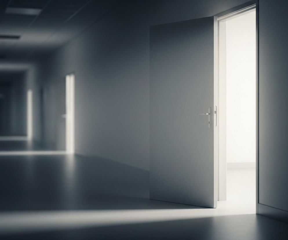Otworzone drzwi ukazują jasne światło we wnętrzu, które kontrastuje z ciemnym, długim korytarzem. Skrzydło drzwiowe jest lekko uchylone w prawo, z widoczną srebrną klamką. Promień światła zza drzwi pada ukośnie na ciemną podłogę korytarza. W oddali korytarza widać kolejne, słabo oświetlone otwory, a cała scena utrzymana jest w chłodnych odcieniach szarości i błękitu.