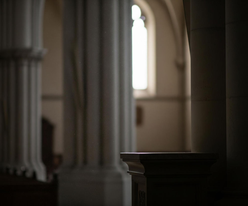 Wnętrze budowli o architekturze sakralnej lub klasycznej, z rzędem filarów i rozświetlonym oknem w tle. Na pierwszym planie, po prawej stronie, widać ciemny, masywny filar oraz jego ozdobną, jaśniejszą podstawę z płaskim blatem. Na środku obrazu widoczne są drobne, rozproszone jasne punkciki, prawdopodobnie kurz, unoszące się w powietrzu, podświetlone promieniami światła wpadającymi z lewej strony. W tle, w głębi, znajduje się rząd smukłych, jasnych kolumn, a dalej wyraźnie widoczne jest wysokie, zaokrąglone u góry okno, z którego wpada intensywne, białe światło.