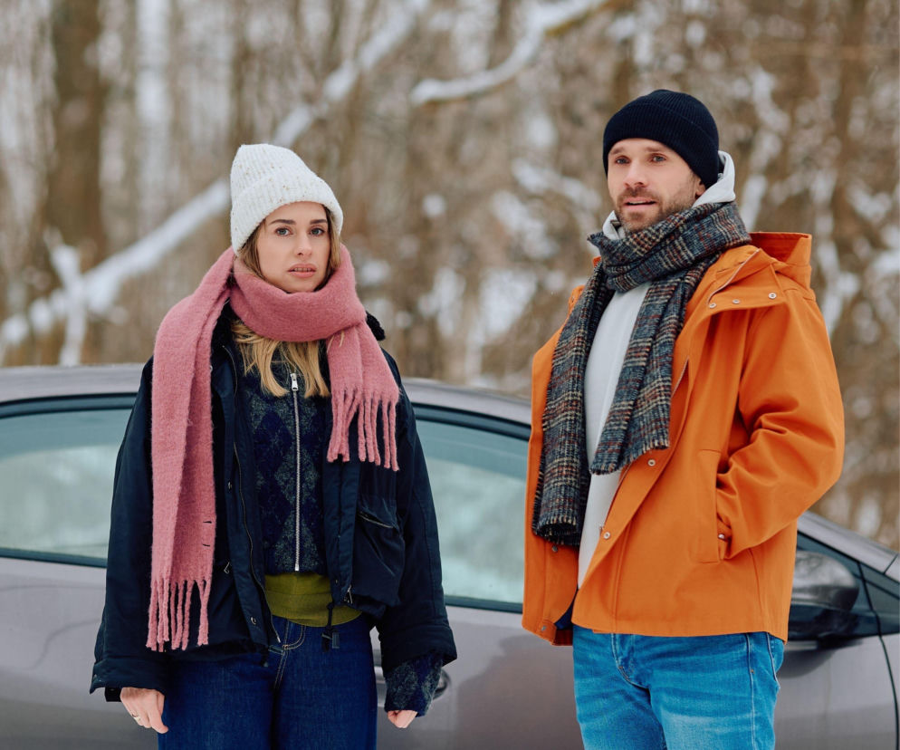 Eliza i Bartek z serialu Na Wspólnej w zimowych stylizacjach, stojący na tle samochodu i ośnieżonego krajobrazu. Ich zaskoczone miny odzwierciedlają dramatyczne wydarzenia z 4203. odcinka. Więcej o zdradzie Elizy przeczytasz na Super Seriale.