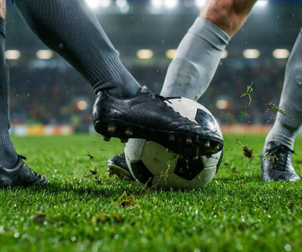 Zbliżenie na buty piłkarskie dwóch zawodników walczących o piłkę na zielonej murawie, z tłem rozmytego stadionu wypełnionego kibicami podczas wieczornego meczu. Scena przedstawia zaciętą rywalizację i dynamikę mistrzostw świata 2026, o których przeczytasz na naszym portalu.