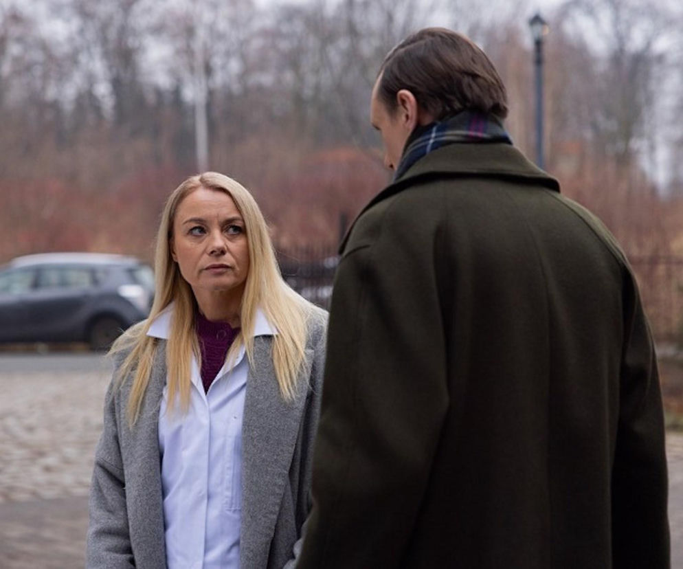 Emilka (Anna Ilczuk) w jasnej koszuli, szarym płaszczu i fioletowym swetrze, z blond włosami, patrzy z powagą na Fryderyka (Andrzej Kłak), który stoi do niej tyłem w ciemnozielonym płaszczu. Scena z serialu Pierwsza miłość podkreśla dramatyzm sytuacji wokół ciąży Emilki. Więcej o ich losach przeczytasz na Super Seriale.