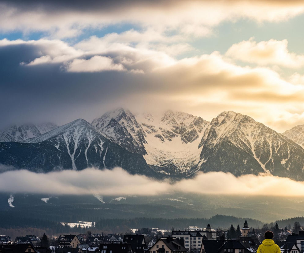 Zasypane śniegiem szczyty górskie dominują na horyzoncie, częściowo przesłonięte chmurami. Lewa strona nieba jest ciemnoszara i niebieska z białymi obłokami, natomiast prawa strona rozjaśnia się do ciepłych, złotych i pomarańczowych tonów. Poniżej gór rozciąga się pasmo ciemnych, iglastych lasów, które stopniowo przechodzi w mglistą warstwę. Na samym dole obrazu widoczne są dachy budynków mieszkalnych, a w prawym dolnym rogu stoi osoba w jaskrawożółtej kurtce, odwrócona plecami do obserwatora.