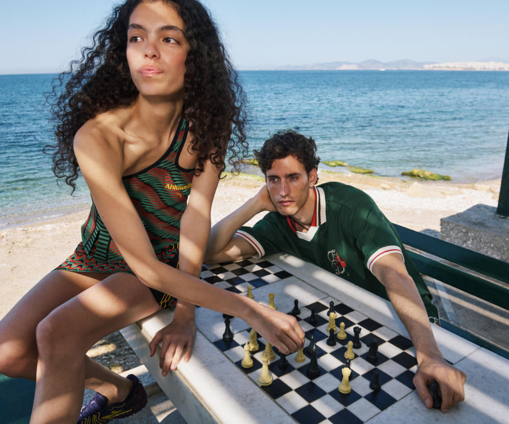 Modelka Julie Garcia i model Ante Padovan grają w szachy na marmurowym stole na plaży w Atenach, prezentując letnią kolekcję PRM. Julie w asymetrycznym topie Ahluwalia, Ante w zielonej koszulce, odzwierciedlają ideę świadomego przeżywania lata, o czym przeczytasz na naszym portalu.