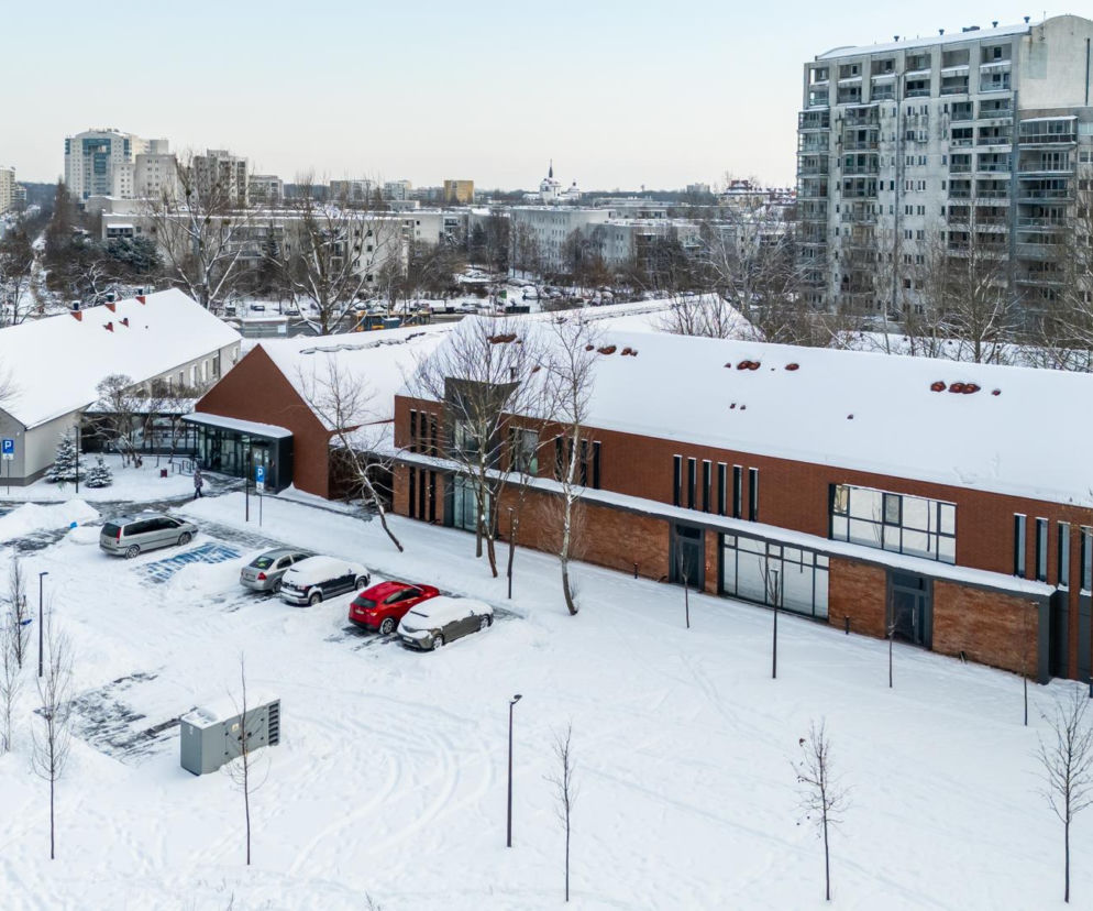 Zimowy widok na Ursynowskie Centrum Opiekuńczo-Mieszkalne przy ul. Belgradzkiej w Warszawie, z budynkami pokrytymi śniegiem, otoczonymi drzewami i ośnieżonym parkingiem. W tle bloki z wielkiej płyty oraz inne budynki mieszkalne. Więcej o architekturze przeczytasz na Architektura Murator Plus.