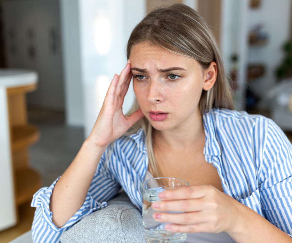 Młoda kobieta z jasnymi włosami trzymająca szklankę wody, z dłonią przy skroni, co sugeruje ból głowy lub zmęczenie, siedząca w domowym wnętrzu. Na Poradnik Zdrowie przeczytasz o tym, jak odwodnienie wpływa na samopoczucie.