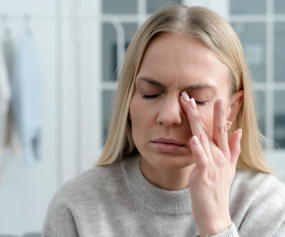 Kobieta w beżowym swetrze pocierająca dłonią powieki, odczuwająca dyskomfort lub zmęczenie oczu. Przedstawia potencjalny objaw syndromu śniegu optycznego, o którym przeczytasz więcej na Poradnik Zdrowie.