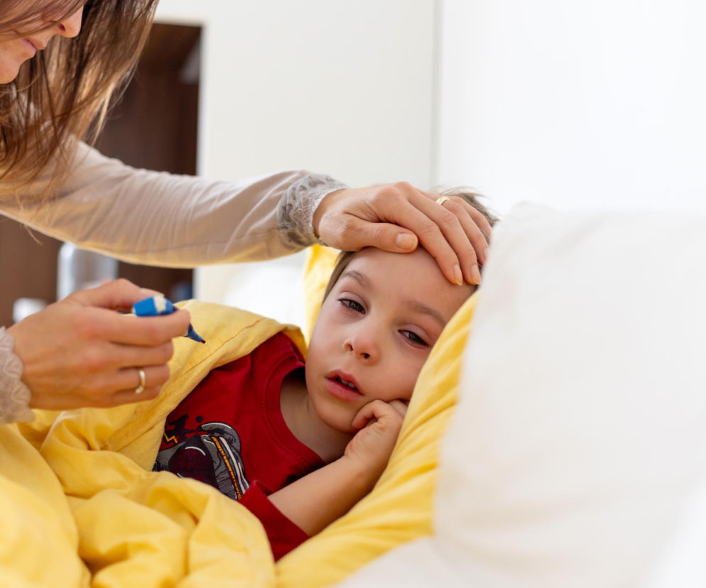 Chore dziecko w łóżku pod żółtą kołdrą z gorączką, matka mierzy mu temperaturę i dotyka czoła, sprawdzając ciepłotę ciała. W artykule Poradnik Zdrowie znajdziesz informacje o supergrypie i jej objawach u dzieci.