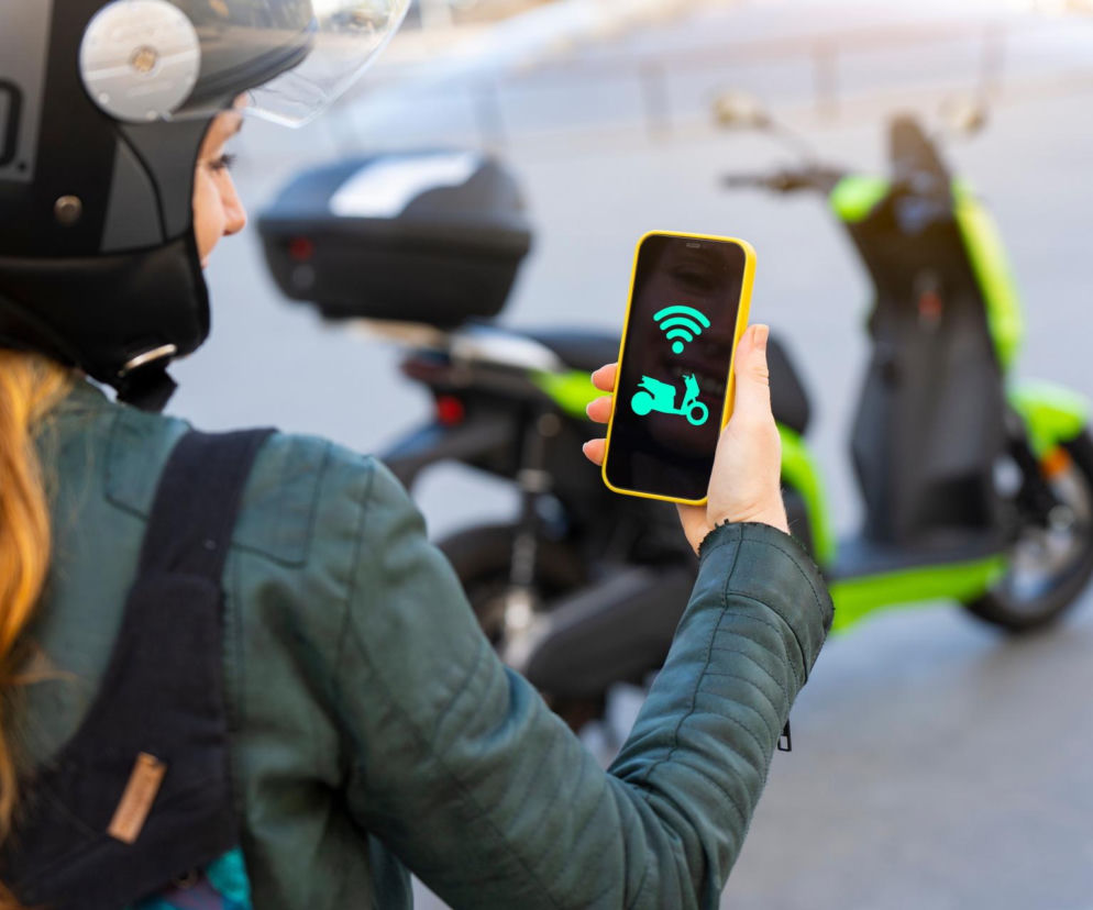 Kobieta w kasku trzyma smartfon z ikoną skutera. W tle stoi zielony skuter. O zmianach dla e-pojazdów czytaj na SE Superbiz.