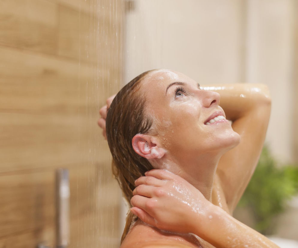 Uśmiechnięta kobieta z zamkniętymi oczami i spienioną skórą pod strumieniem wody w kabinie prysznicowej, co symbolizuje relaks i higienę, ale w kontekście artykułu z portalu Poradnik Zdrowie przestrzega przed szkodliwym nawykiem sikania pod prysznicem.