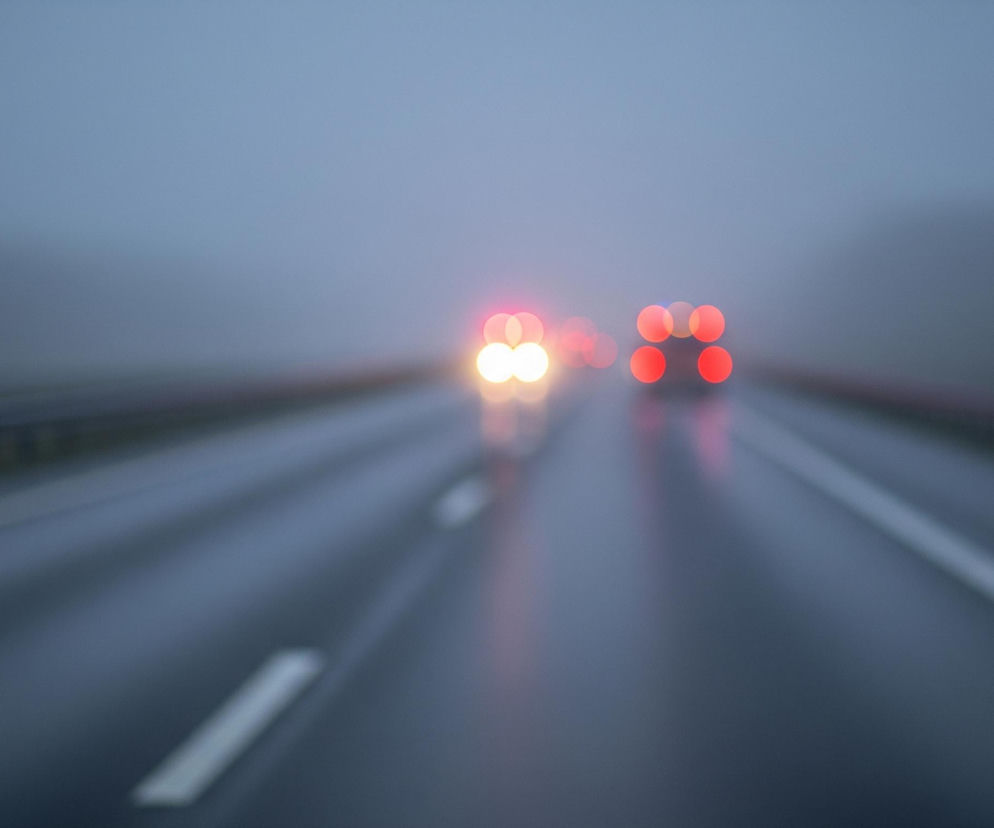 Scena przedstawia rozmazaną perspektywę drogi, gdzie dominują stonowane odcienie szarości i niebieskiego. W oddali widoczne są rozmyte światła samochodów: po lewej stronie jasne, biało-żółte reflektory, a po prawej wyraźnie czerwone światła tylne, wyglądające jak cztery okręgi. Powierzchnia jezdni jest mokra i błyszcząca, z widocznymi refleksami świateł, a linia przerywana na środku drogi, w lewej części kadru, jest lekko ukośna. Otoczenie jest zamglone, niewyraźne, z ledwo zarysowanymi sylwetkami drzew lub konstrukcji po prawej stronie.