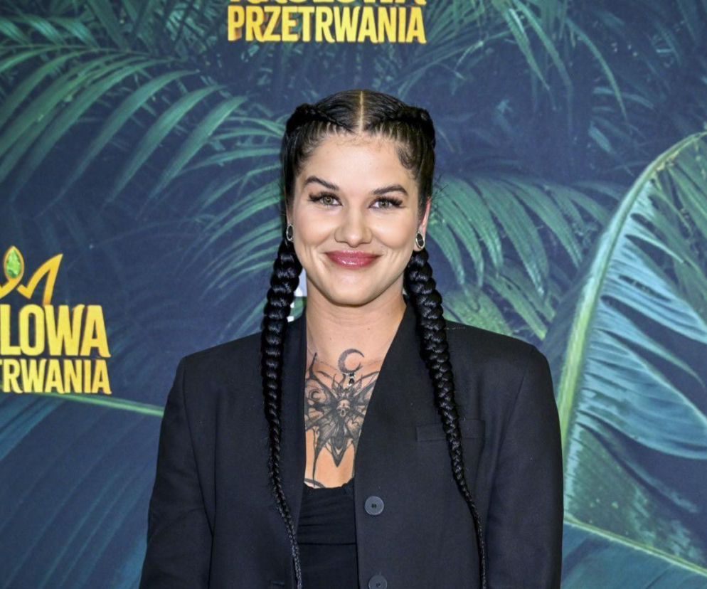 Dominika Rybak, zwyciężczyni Królowej Przetrwania, uśmiecha się na tle baneru programu. Ma czarne włosy splecione w warkocze i widoczny tatuaż na mostku. Artykuł o jej walce z chorobą SIBO przeczytasz na naszym portalu.