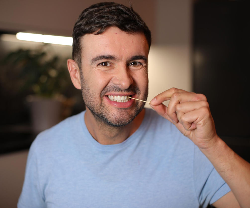 Mężczyzna w niebieskiej koszulce z uśmiechem używający wykałaczki do czyszczenia zębów. To nawyk, który może uszkodzić dziąsła i szkliwo, zwiększając ryzyko chorób serca, o czym przeczytasz więcej na Poradnik Zdrowie.