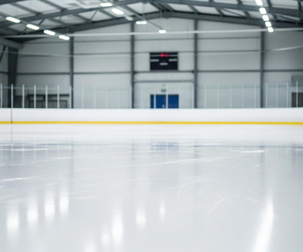 Lodowisko hokejowe z gładkim, białym lodem pokrytym licznymi, delikatnymi zadrapaniami i śladami po łyżwach dominuje w kadrze. Na pierwszym planie, w prawej dolnej części obrazu, wzdłuż żółtej linii bandy, ustawione są czarne łyżwy hokejowe z białymi sznurowadłami i ostrzami, sugerując gotowość do użycia. Bandy, oddzielające lód od widowni, są białe u dołu i ciemnoniebieskie u góry, z żółtą linią na ich styku. W tle widać puste trybuny oraz tablicę wyników z czerwonymi cyframi, a także konstrukcję hali z rzędami jasnych lamp sufitowych.