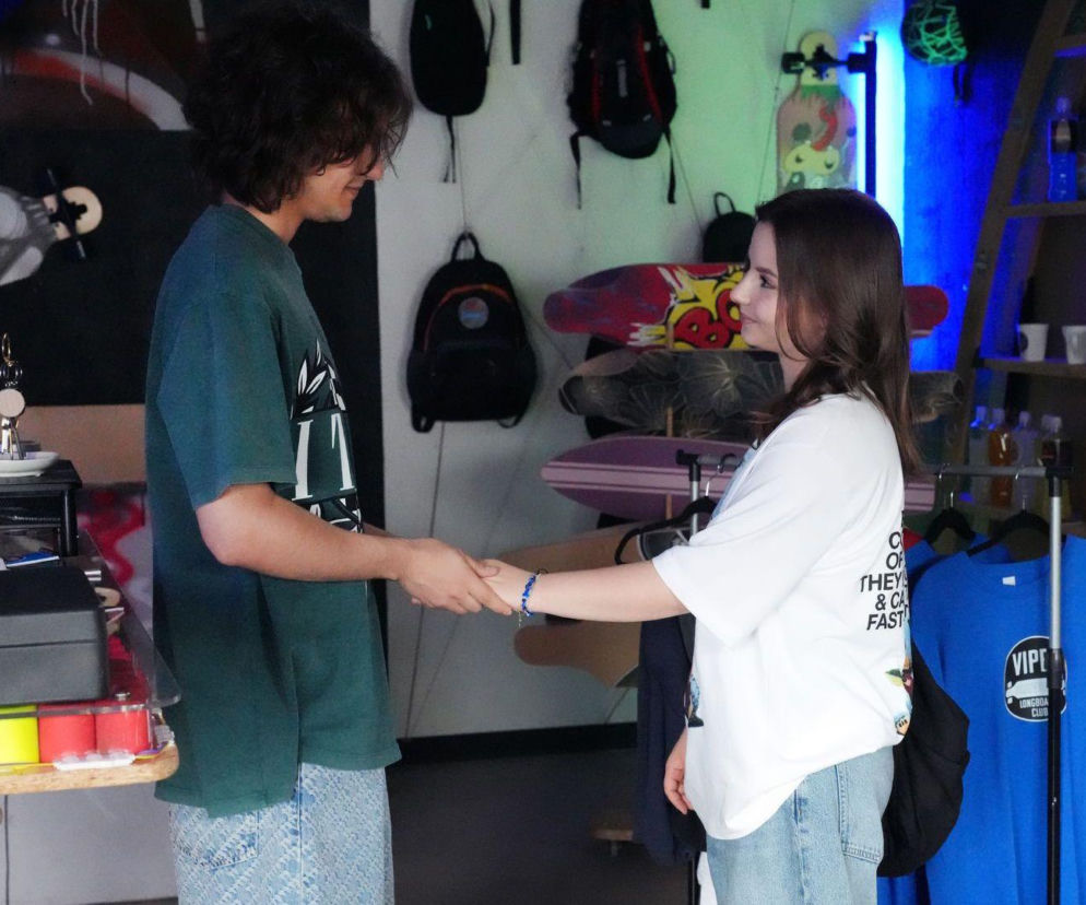 Młody mężczyzna i kobieta trzymają się za ręce, stojąc naprzeciwko siebie w skateshopie, patrząc na siebie z uśmiechem. Scena z serialu Barwy szczęścia, gdzie Romeo wyznaje Ewie miłość. Więcej o ich losach na Super Seriale.