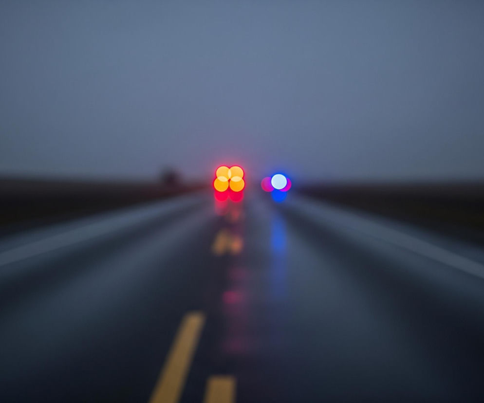 Droga, ciemnoszara i mokra od deszczu, rozciąga się w głąb obrazu, gdzie na horyzoncie zlewają się ze sobą niebo i płaski, ciemny krajobraz. Na asfalcie widoczne są dwie przerywane linie, żółta po lewej i biała po prawej, prowadzące perspektywicznie ku górze. W oddali, w centrum kadru, wyróżniają się jaskrawe, rozmyte światła: cztery czerwone lub pomarańczowe po lewej i dwa niebieskie po prawej, odbijające się delikatnie na mokrej nawierzchni. Całość jest nieostra, tworząc efekt bokeh, z dominującymi odcieniami szarości i granatu, przełamanymi jedynie intensywnymi barwami świateł.